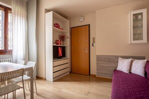 Ein Zimmer mit Holzboden, einem Bett mit violetter Bettdecke, einem Fernseher auf einem Regal, einer Holztür und einem Fenster mit Vorhängen.
