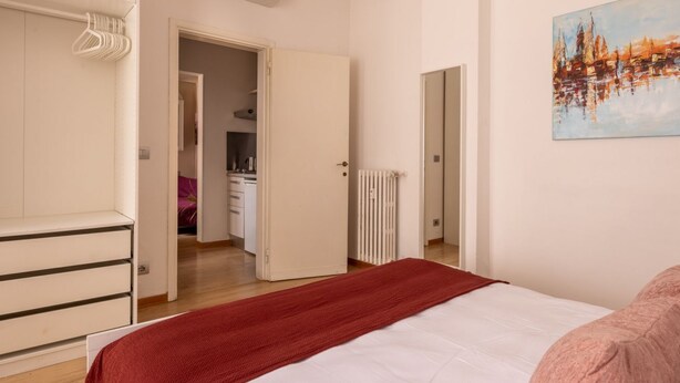 Un dormitorio con cama, una manta roja, un radiador blanco y un cuadro en la pared.