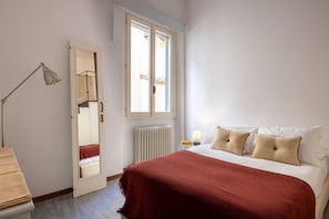 Ein Schlafzimmer mit Bett, Nachttischlampe, Spiegel, Fenster und Heizkörper.