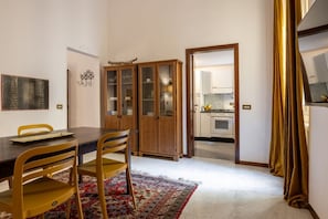 Ein Essbereich mit Holztisch, Stühlen und einem gemusterten Teppich. Ein Schrank mit Glastüren und eine Küche sind durch einen offenen Durchgang sichtbar.