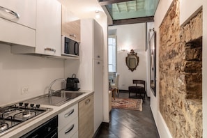 A modern kitchen with a gas stove, a microwave, and a sink. There is a dining area with a chair and a rug. A stone wall and a framed mirror are visible. - Calimaruzza Apartment - by Maison Florence (Firenze)
