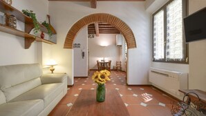Un salon comprenant un canapé, une table basse en bois sur laquelle est posé un vase rempli de tournesols, un téléviseur et une fenêtre ornée de rideaux à motifs.