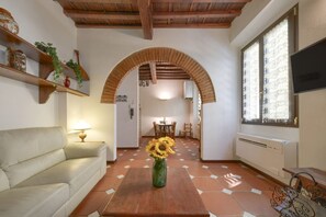 Ein Wohnzimmer mit einer Couch, einem Holztisch mit einer Vase voller Sonnenblumen, einem Fernseher und einem Fenster mit gemusterten Vorhängen.