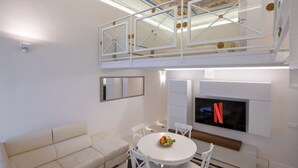 Uma sala de estar moderna com sofá branco, mesa de jantar redonda com cadeiras, televisão na parede e decoração no teto.