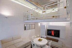 A modern living room with a white sofa, a round dining table with chairs, a TV mounted on the wall, and a decorative ceiling. - The White House - Home of the Frescoes (Firenze)