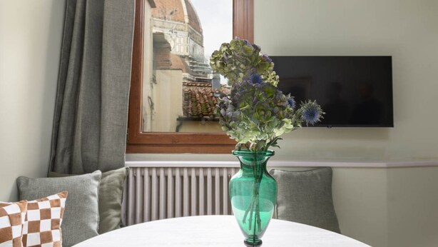 Una habitación con una mesa redonda, un florero verde con flores, un televisor en la pared y una ventana con vistas a un edificio.