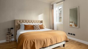 Un dormitorio con cabecera acolchada, cama grande con colcha marrón, mesita de noche, espejo y ventana con cortinas.