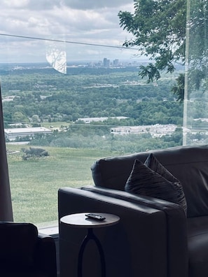 A room with a view of a green landscape, a leather sofa, and a small round table with a remote control. - Welcome to Skyline Point ~ Louisville Skyline (Floyds Knobs)
