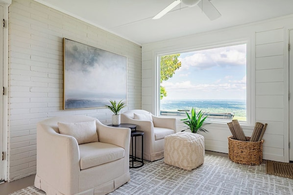 A room with a white brick wall, a large painting, two sofas, a small table, a potted plant, a wicker basket, and a view of the ocean through a window.
