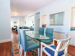 Ein Essbereich mit einem Glastisch und blauen Stühlen, ein weißer Stuhl mit blauer Sitzkissen sowie eine Heizung und ein Spiegel an der Wand.