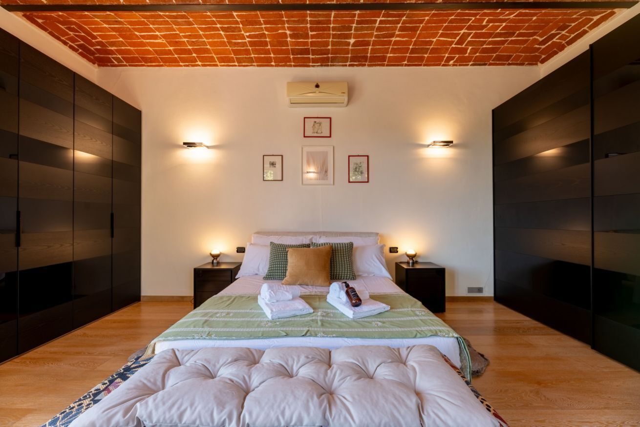 A bedroom with a brick ceiling, a bed with a green and white bedspread, two bedside tables with lamps, and framed pictures on the wall.
