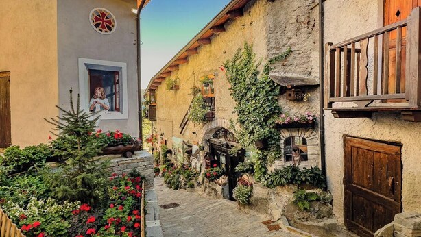 Una estrecha calle empedrada flanqueada por edificios, un balcón de madera y un pequeño jardín con flores.