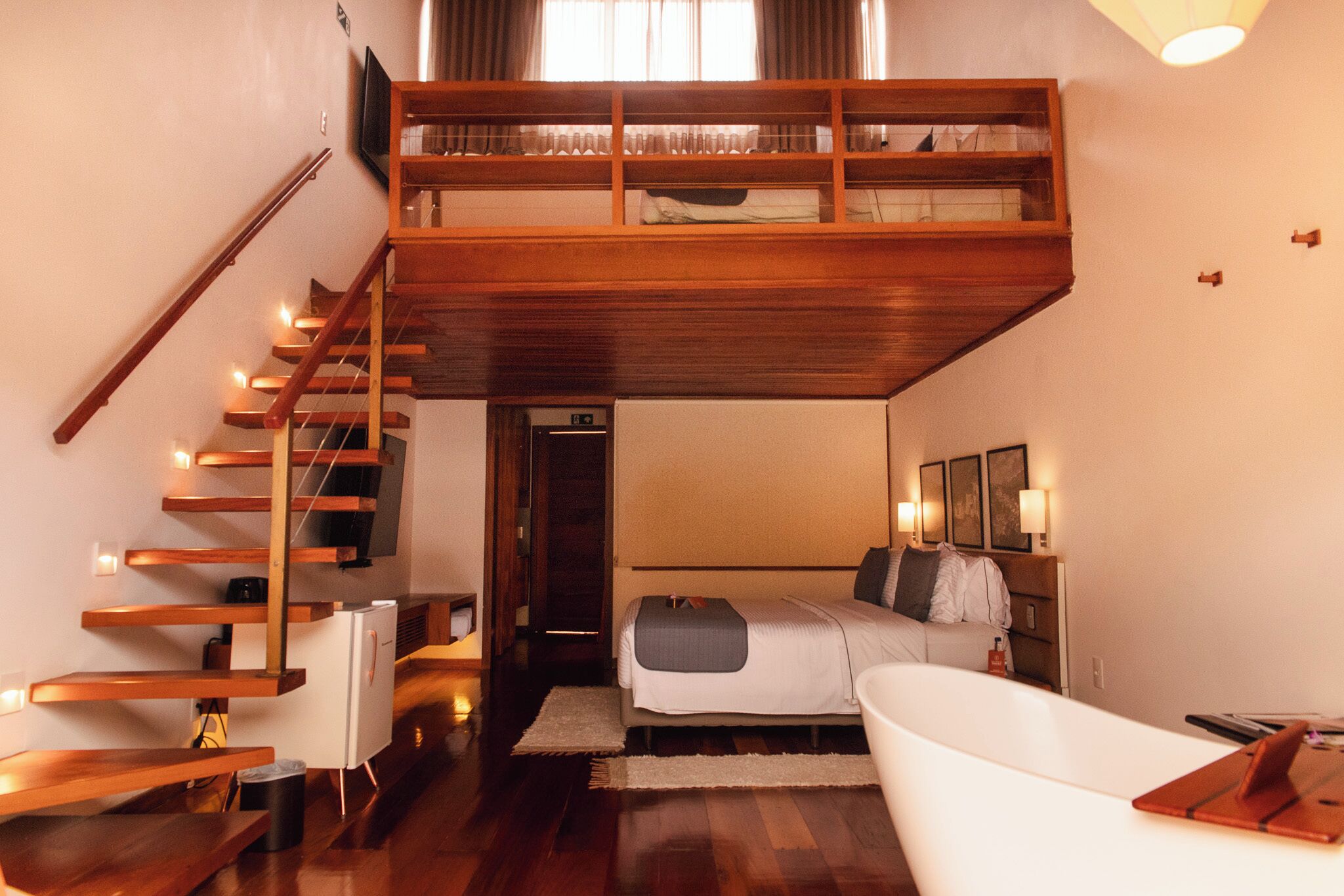 A loft-style bedroom with a wooden staircase, a bed, a white bathtub, and a flat-screen TV.