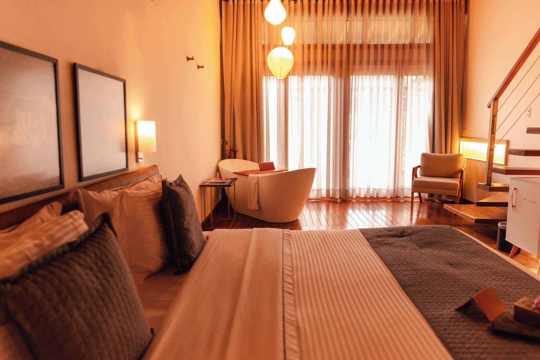 Quarto de hotel com cama, sofá, poltrona, escrivaninha e uma janela grande com cortinas.