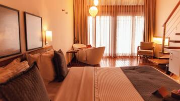 Quarto de hotel com cama, sofá, poltrona, escrivaninha e uma janela grande com cortinas.