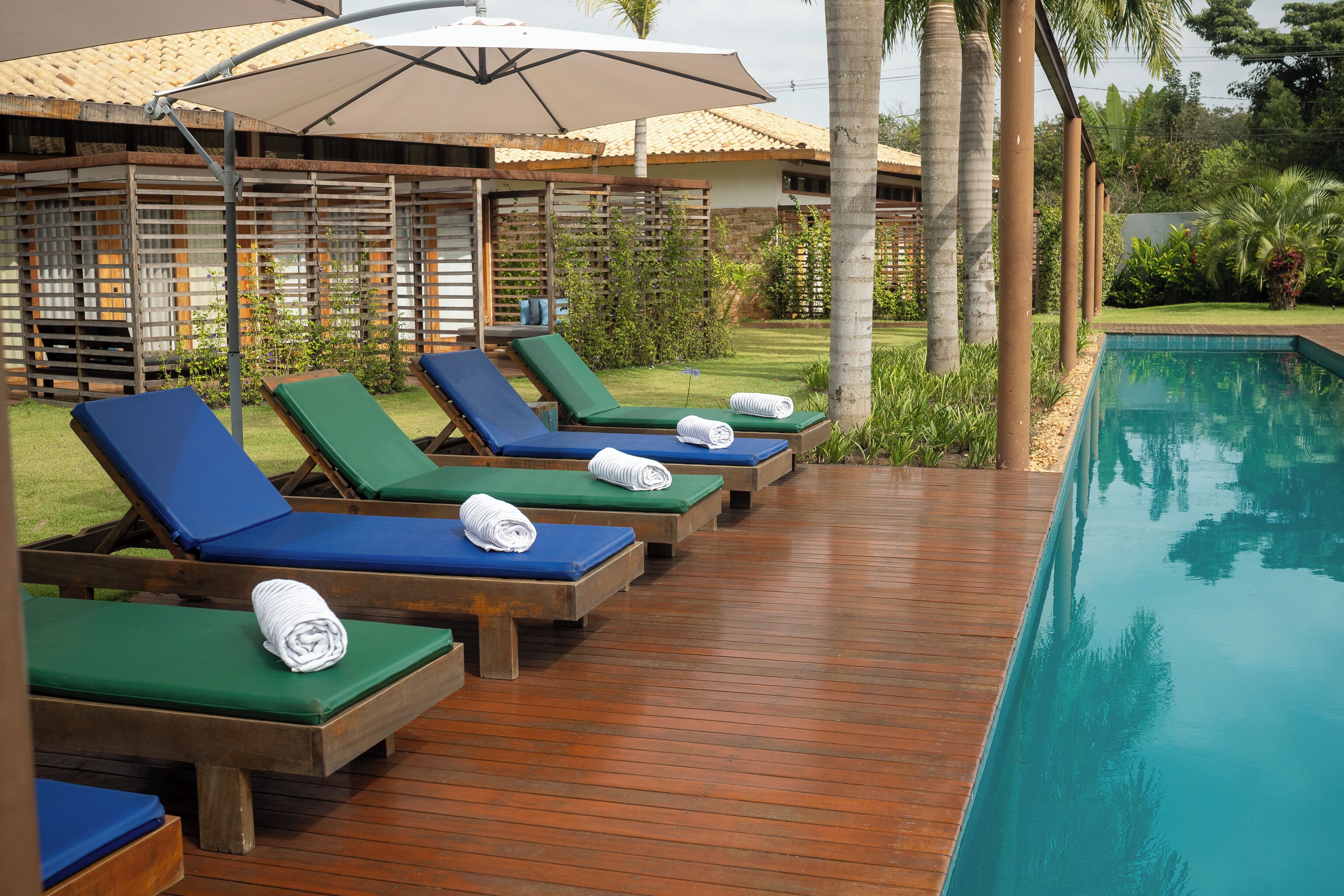 A poolside area with lounge chairs and umbrellas, surrounded by greenery and a building with a tiled roof.