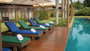 A poolside area with lounge chairs and umbrellas, surrounded by greenery and a building with a tiled roof.