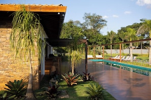 Un área junto a la piscina con un edificio de piedra, sillas de descanso y plantas tropicales.