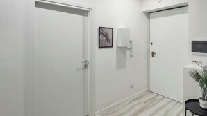 A hallway with two white doors, a framed picture on the wall, a small table with a plant, and a microwave on a white counter. - [Comfort Suite] Airport 5 min • A/C • Smart TV (Genova)