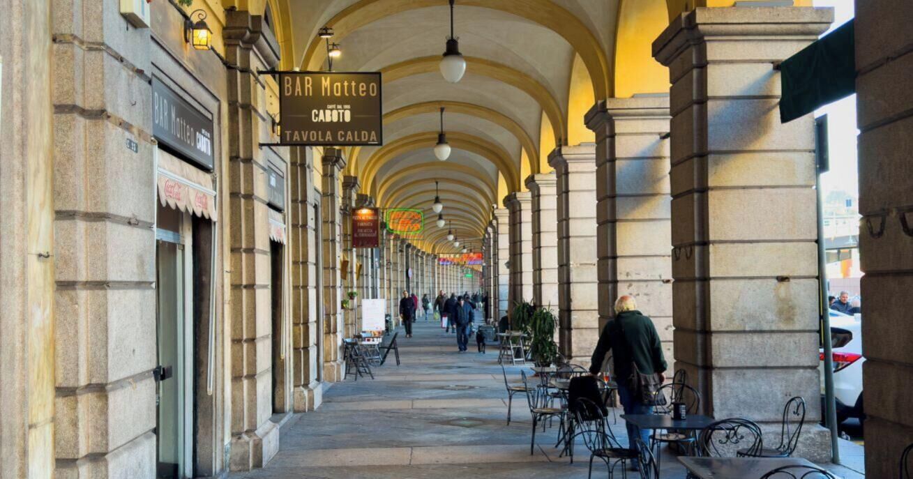 Ein überdachter Fußweg mit Bögen, Außenbestuhlung und Schildern für eine Bar und ein Restaurant.
