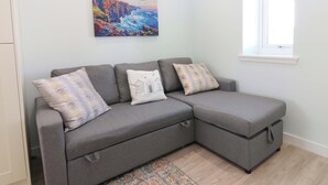 Ein graues Ecksofa mit drei Kissen, ein farbenfrohes Gemälde an der Wand und ein Fenster mit einer beigen Jalousie.
