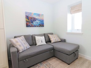 Ein graues Ecksofa mit drei Kissen, ein farbenfrohes Gemälde an der Wand und ein Fenster mit einer beigen Jalousie.
