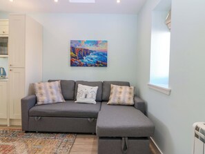 A grey sofa with three pillows, a colorful painting on the wall, and a window with blinds. - Lightkeepers Bothy (Eyemouth)