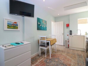 A room with a TV, a painting, a chair, a table, a rug, and a kitchen area. - Lightkeepers Bothy (Eyemouth)