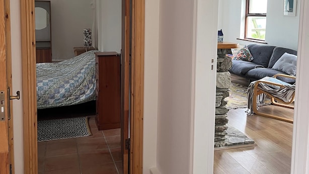 A room with a bed, a wooden door, a stone wall, and a view of a living area with a sofa and a window.