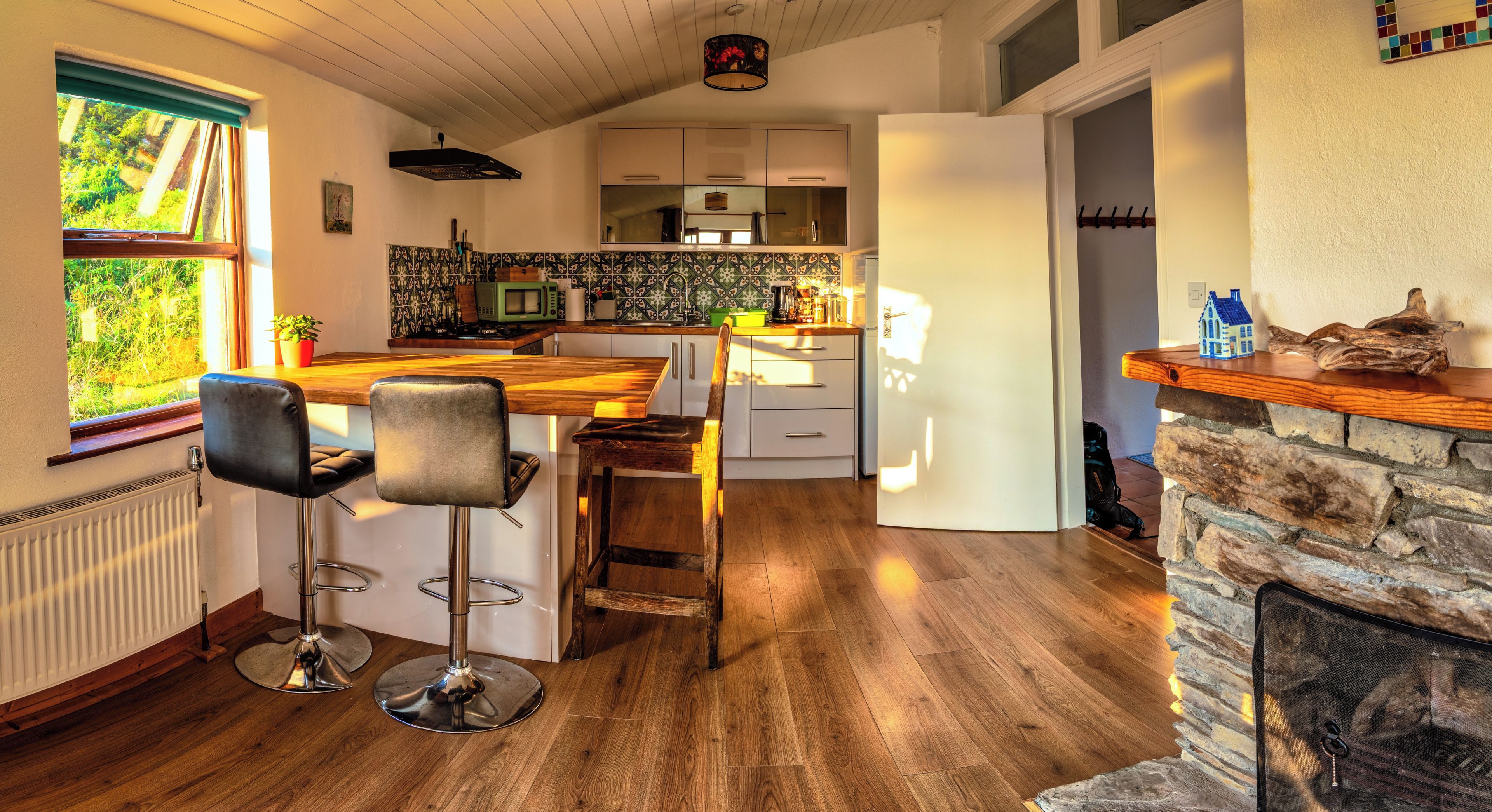 A kitchen with a wooden island, bar stools, a stone fireplace, and a view of greenery outside.