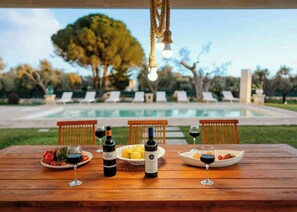 Ein Holztisch mit Weinflaschen, Gläsern und Tellern voller Essen, mit Blick auf einen Swimmingpool und Liegestühle.