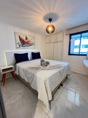 Quarto com cama, mesa de cabeceira, pintura, guarda-roupa e janela.