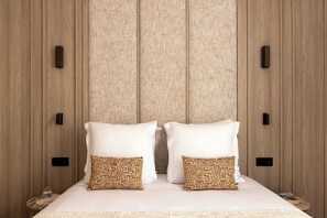 Ein Bett mit weißen Leinen und gemusterten Kissen, ein Holz-Kopfschild und ein Nachttisch mit einem Glas Wasser.