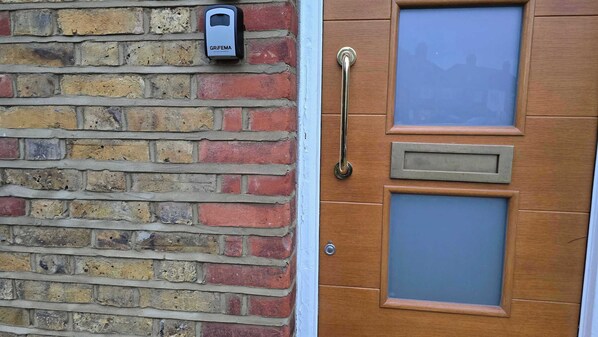 A wooden door with a metal handle, a glass panel, and a GRIFEMA device mounted on the brick wall. - Spacious Family Home 4 Beds Garden Parking (Greater London)