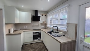 A compact kitchen with a built-in oven, a sink with a dish rack, and a window with blinds. - Spacious Family Home 4 Beds Garden Parking (Greater London)