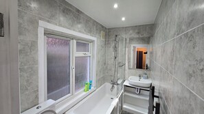 A modern bathroom with a bathtub, a window with frosted glass, and a sink with a mirror above it. - Spacious Family Home 4 Beds Garden Parking (Greater London)