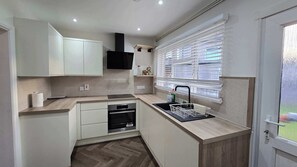 A compact kitchen with a built-in dishwasher, a window with blinds, and a door leading to an outdoor area. - Spacious Family Home 4 Beds Garden Parking (Greater London)