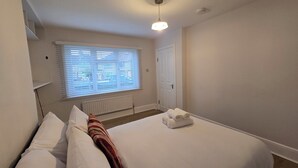 Ein Schlafzimmer mit einem Bett, einem Heizkörper, einem Fenster mit Jalousien, einer Tür und einer Deckenleuchte.