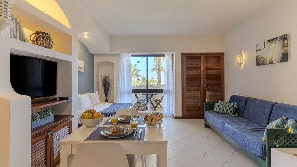 Una sala de estar con sofá, mesa de comedor, televisor y vista a palmeras a través del ventanal.