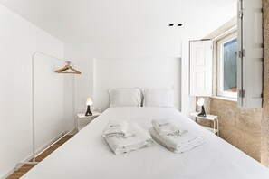 Ein schlicht eingerichtetes Hotelzimmer mit einem großen Bett, zwei Nachttischen mit Lampen, einem Holzaufhänger und einem Fenster mit geöffneten Fensterläden.