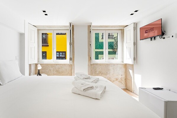 A hotel room with a bed, a TV, and two windows with views of buildings. - GuestReady - InPorto Building - Room 3 (Porto)