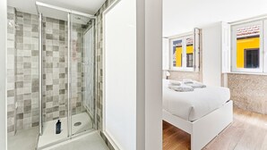 Un baño moderno con ducha envuelta en cristal, cama con toallas ordenadamente dobladas y ventana con marco amarillo.