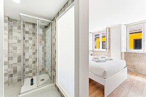 Ein modernes Badezimmer mit einer gläsernen Duschkabine, einem Bett mit ordentlich gefalteten Handtüchern und einem Fenster mit gelbem Rahmen.