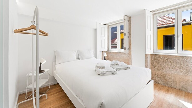 Un dormitorio con cama, perchero, una silla, escritorio y ventana con vistas a edificios.