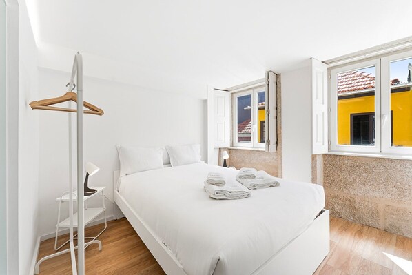 Ein Schlafzimmer mit einem Bett, einem Kleiderhaken, einem Stuhl, einem Schreibtisch und einem Fenster mit Blick auf Gebäude.