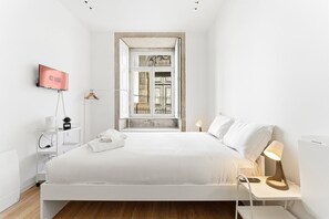 A modern hotel room with a large bed, a TV, a bedside table with a lamp, and a window with a view of a building. - GuestReady - InPorto Building - Room 1 (Porto)