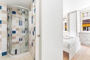 A modern bathroom with a glass shower enclosure, a white sink, and a window with a view of the city. - GuestReady - InPorto Building - Room 2 (Porto)