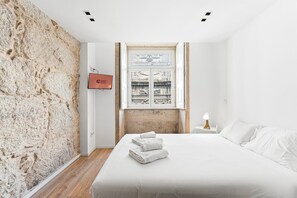 Um quarto com parede de pedra, uma cama com roupa de cama branca, uma mesa de cabeceira com abajur e uma TV na parede exibindo 'GUEST READY'.