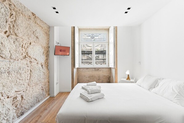 Ein Schlafzimmer mit Steinwand, einem Bett mit weißer Bettwäsche, einem Nachttisch mit Lampe und einem an der Wand befestigten Fernseher, der 'GUEST READY' anzeigt.