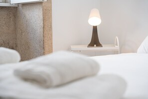 Uma cama bem arrumada com edredom branco, uma mesa de cabeceira com abajur e uma parede com textura.